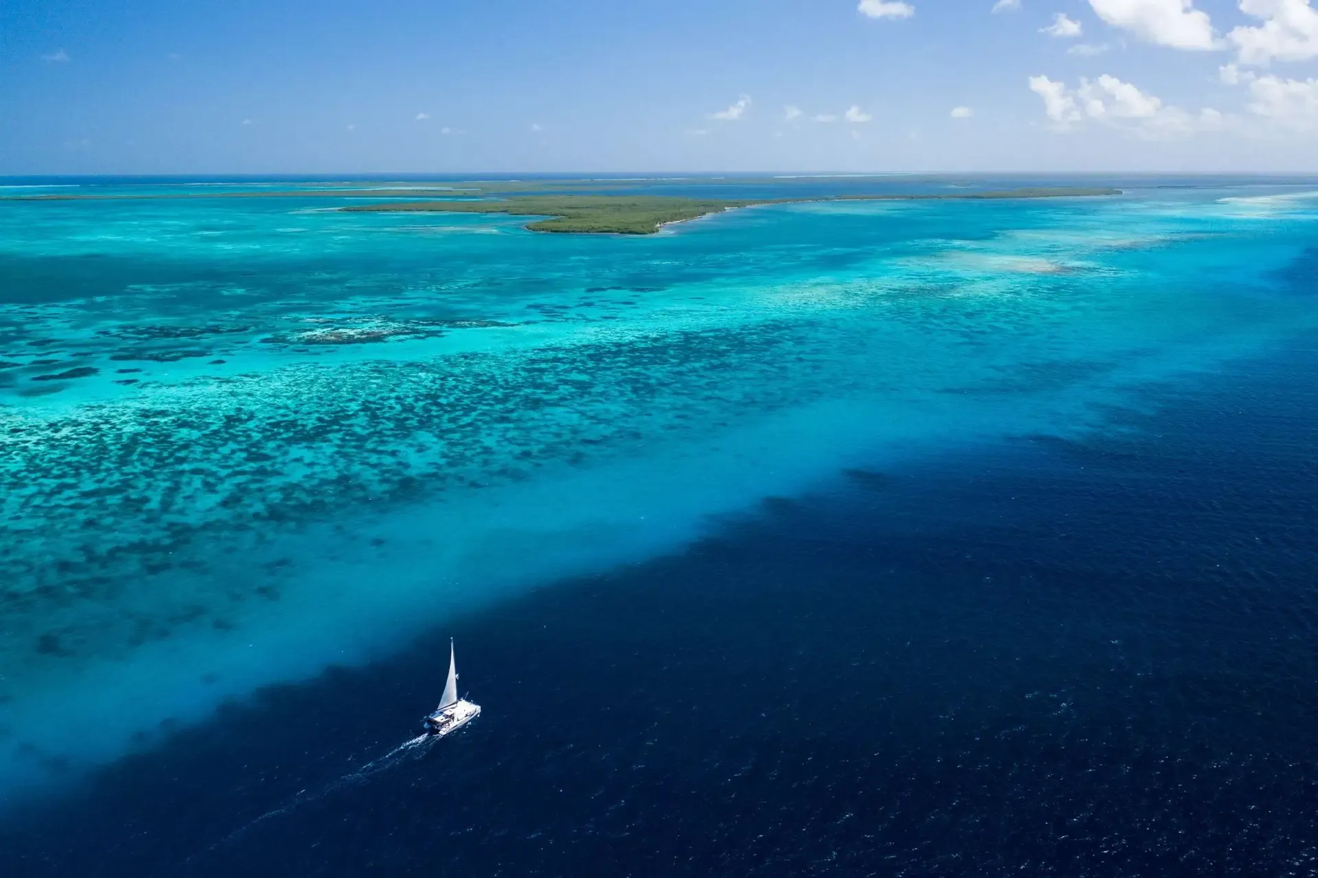 Aerial view of Belize's barrier reef, turquoise flats meeting deep blue water, with a fishing skiff near the reef edge