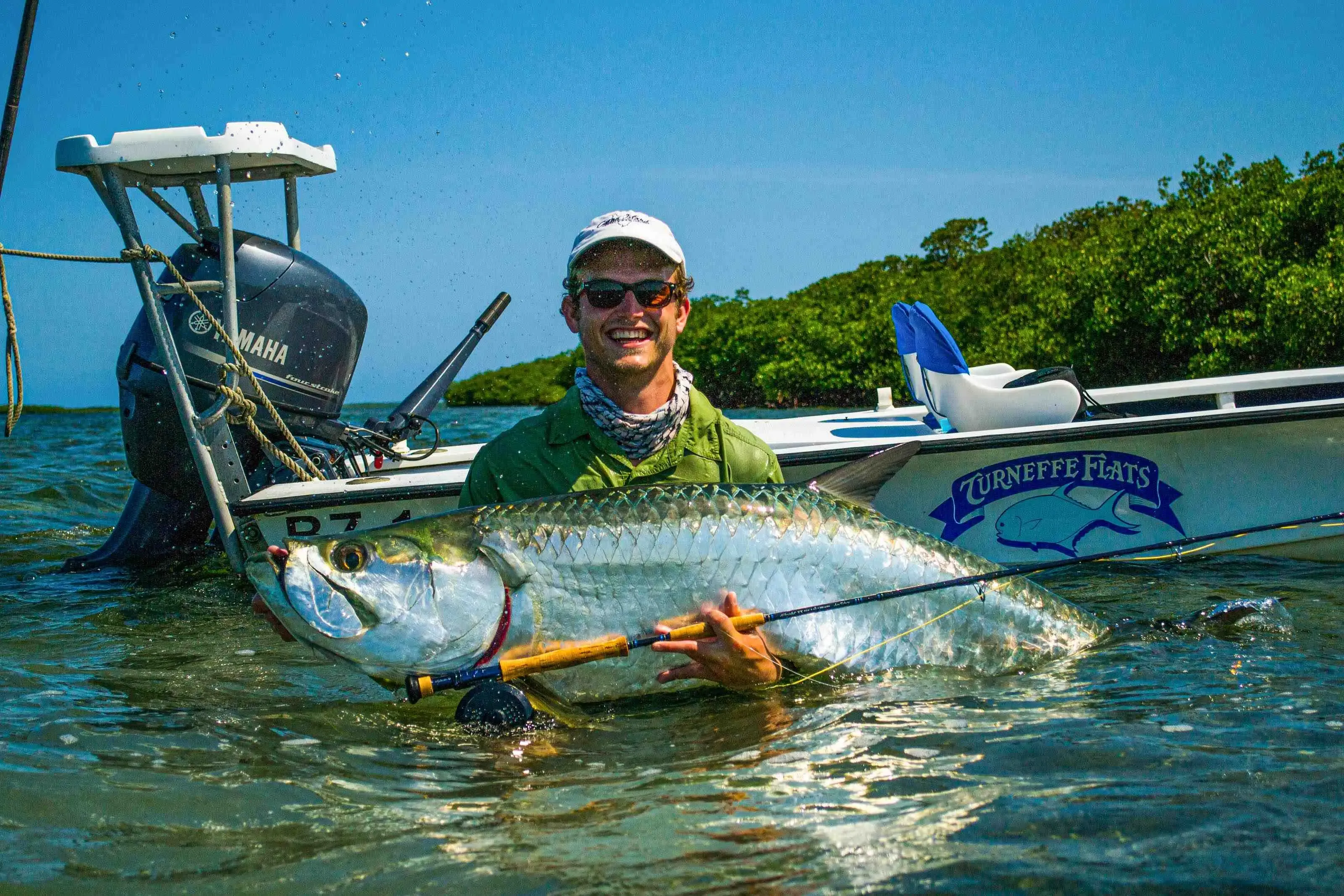 Fly angler mid-cast to tailing permit on a shallow Belizean flat, guide poling the skiff at the stern, early morning light over the water